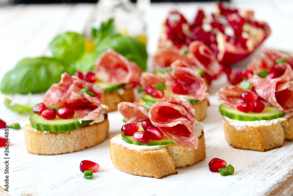 Canape or crostini with toasted baguette, light cheese, cucumber, pomegranate and salami on light background. Top view with copy space.