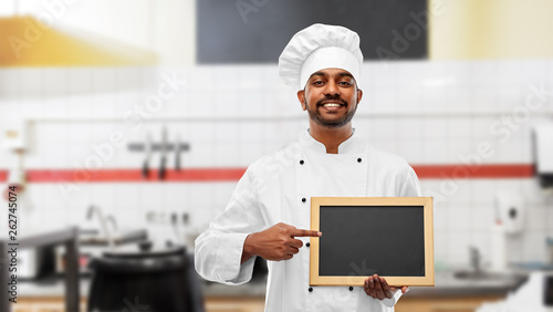 cooking, profession and people concept - happy male indian chef in toque with blank chalkboard for menu over restaurant kitchen background photo
