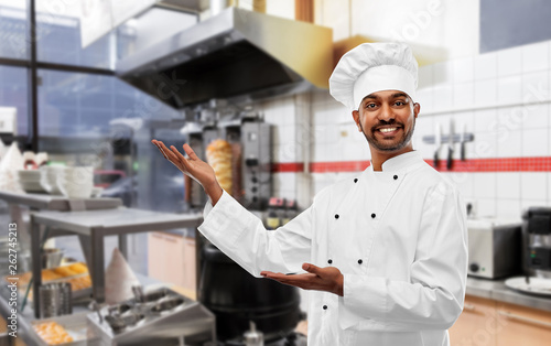 cooking, profession and people concept - happy male indian chef in toque presenting something over kebab shop kitchen background