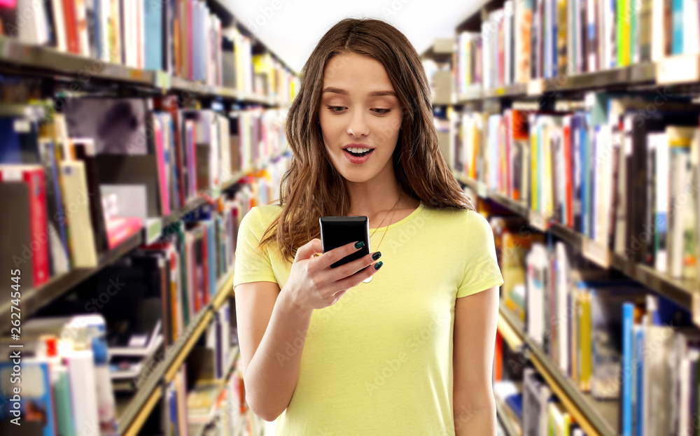 technology, education and people concept - smiling young woman or teenage girl in blank yellow t-shirt using smartphone over book shelves in library background technology, education and people concept - smiling young woman or teenage girl in blank yellow t-shirt using smartphone over book shelves in library background