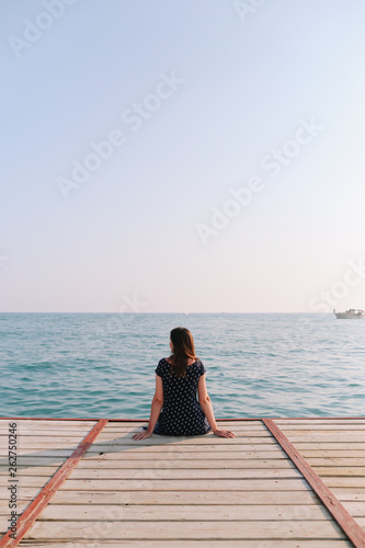 Girl sitting on the pier and looking at the sea horizon and blue sky. Vacation at sea. Summer time sea vacation background 