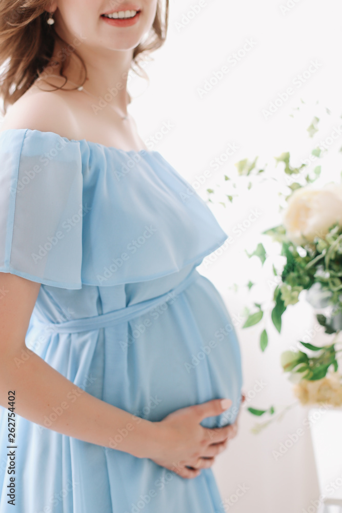 Pregnant woman in blue dress holds hands on belly on a white background. Pregnancy, maternity, preparation and expectation concept. Close-up, indoors. Beautiful tender mood photo of pregnancy. Pregnant woman in blue dress holds hands on belly on a white background. Pregnancy, maternity, preparation and expectation concept. Close-up, indoors. Beautiful tender mood photo of pregnancy.