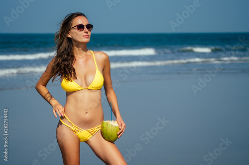 beautiful tanned girl in trendy yellow bathing suit sunglasses stands with coconut in her hands on beach tropical island vacation travel swimwear new season.derox diet idea