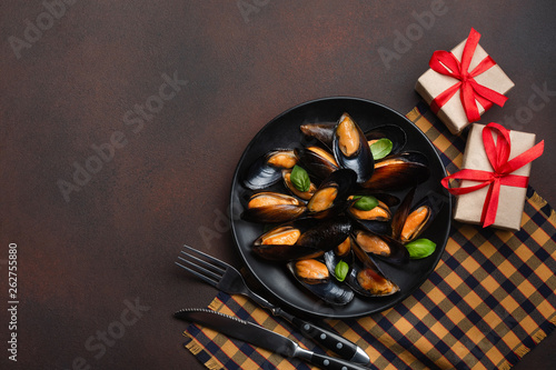 Seafood mussels and basil leaves in a black plate with fork, knife and gift boxes on towel and rusty background