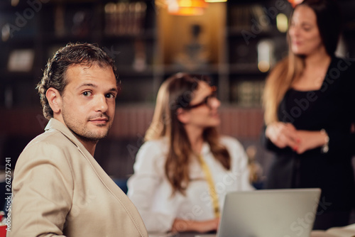 Young arabian businessman dressed smart casual sitting in coffee shop on meeting and looking over shoulder at camera. In background his colleagues discussing about project.