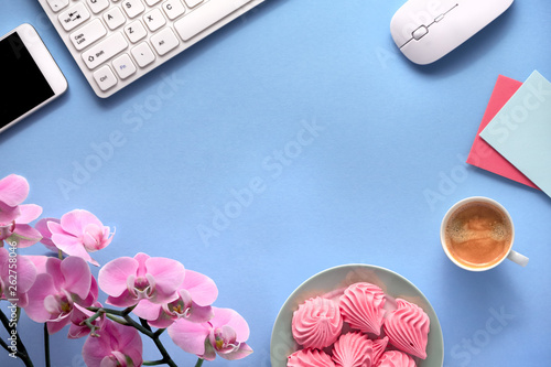 Feminine working space with pink orchids, keyboard, mobile phone, plate of marshmallow and cup of coffee