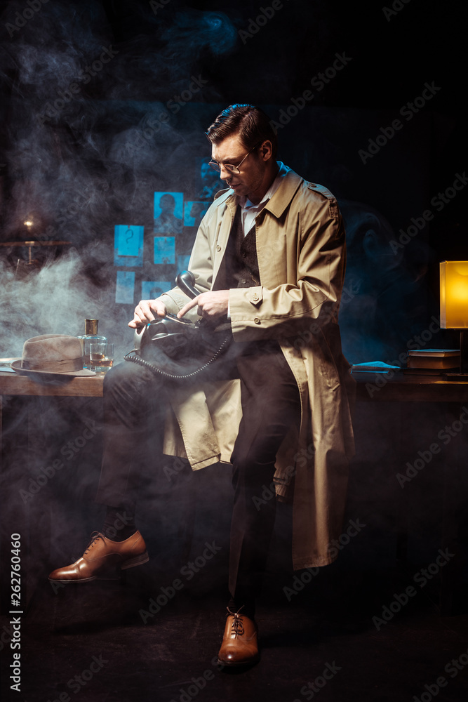 Detective in trench coat sitting on table and using telephone in dark office
