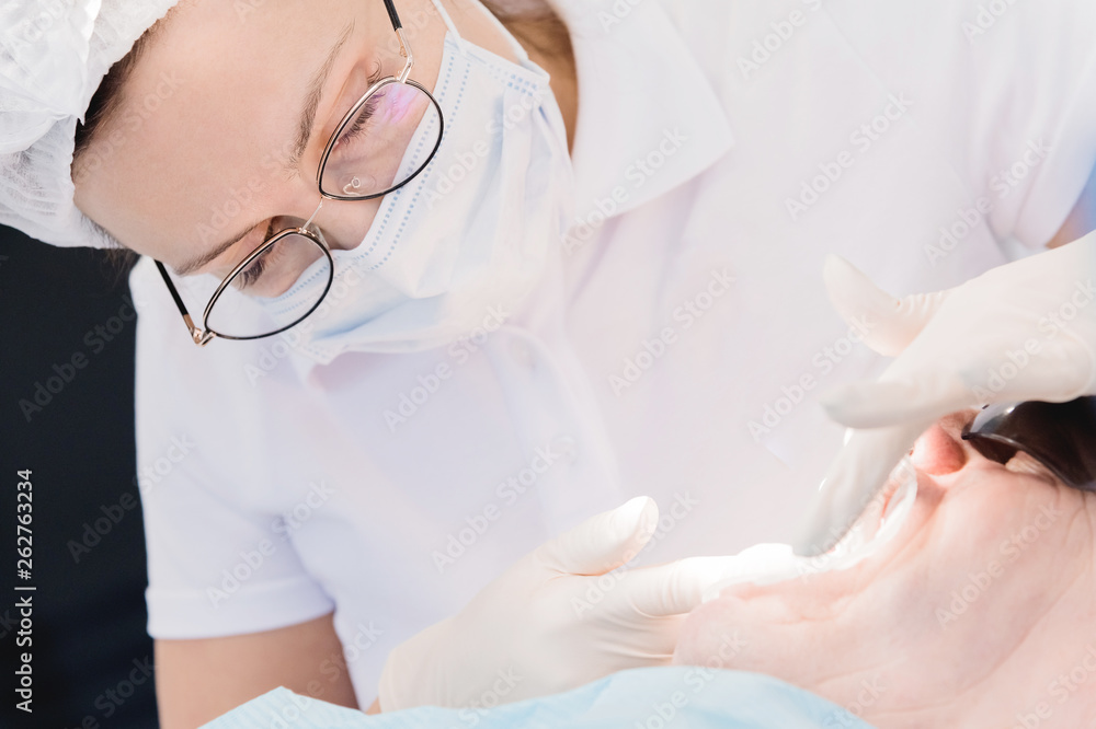 Fototapeta premium Female dentist with an assistant examines the patient's mouth of a man of an aged man. High key professional work of a dentist in the dental clinic