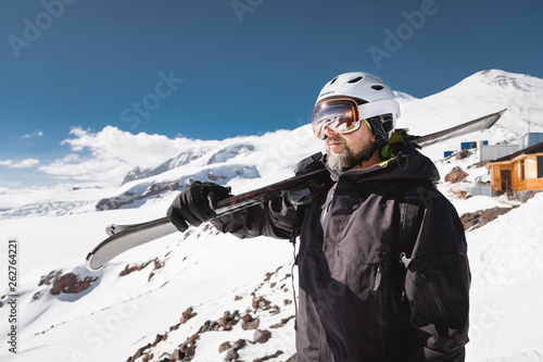 Portrait bearded male skier aged against background of snow-capped Caucasus mountains. An adult man wearing ski googles mask and helmet skis on his shoulder looks mountains. Ski resort concept photo