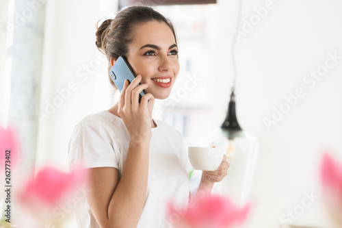 Beautiful young student girl drinking coffee posing indoors talking by mobile phone.
