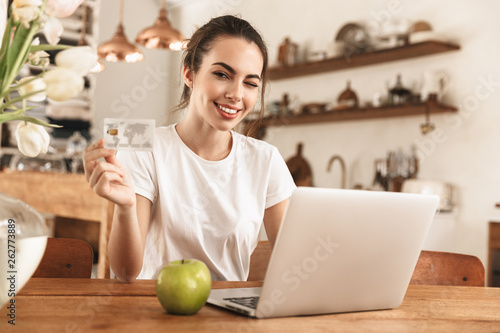 Beautiful young student girl with apple indoors using laptop computer holding credit card.