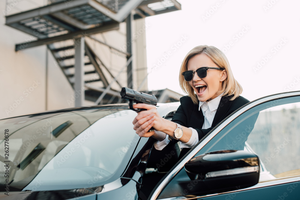 Obraz premium cheerful blonde woman in sunglasses holding gun near black car