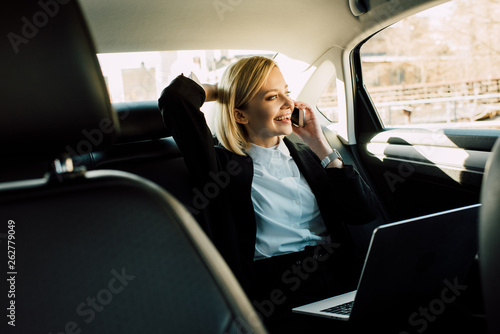 smiling blonde woman talking on smartphone near laptop in car