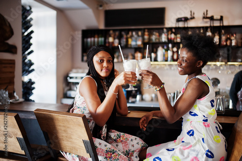 Two black african girlfriends at summer dresses drinking and cheering milkshake cocktails in bar.