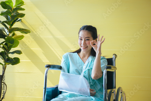 Asian beautiful female patient raise up your hands OK sign symbol and sitting on wheelchair at hospital,Happy and smiling