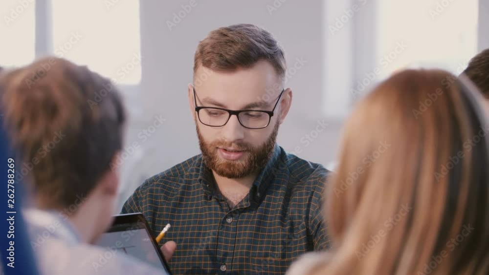 Medium shot of young professional Caucasian businessman talking to colleagues behind table at brainstorm team meeting. Medium shot of young professional Caucasian businessman talking to colleagues behind table at brainstorm team meeting.