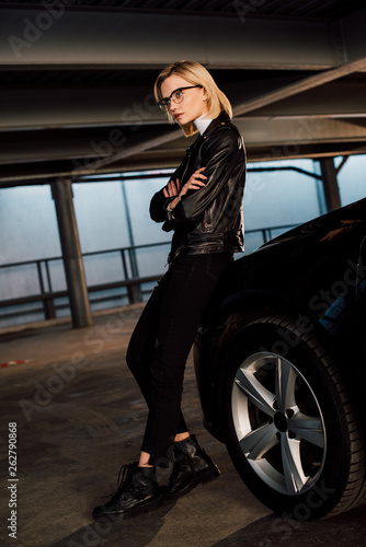 confident young woman in glasses standing in parking with crossed arms near black car © LIGHTFIELD STUDIOS