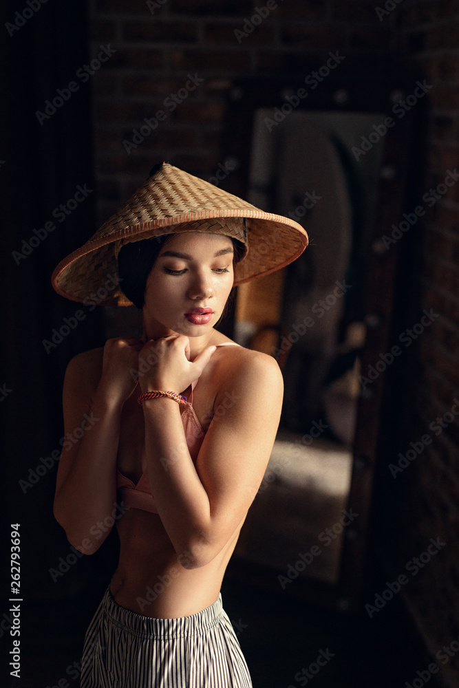 beautiful fit woman with brunette hair in pink bra posing with asian style hat on dark background in studio beautiful fit woman with brunette hair in pink bra posing with asian style hat on dark background in studio