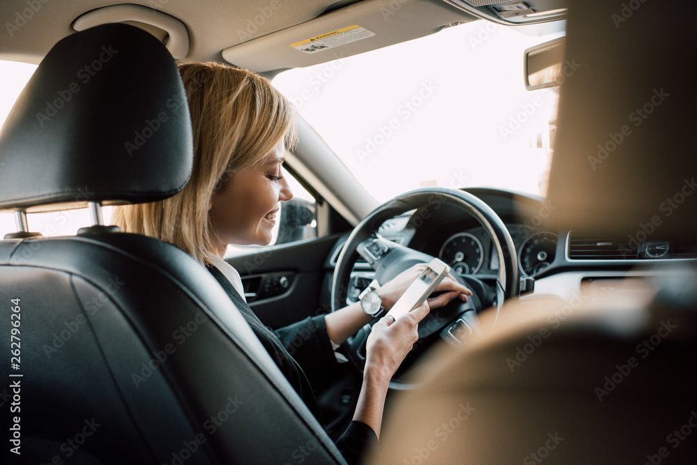 cheerful blonde young woman sitting in car and using smartphone