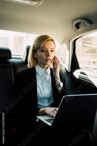 pensive blonde woman talking on smartphone near laptop in car