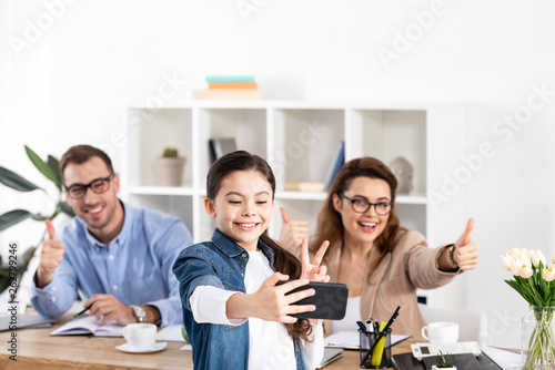 selective focus of cheerful child taking selfie on smartphone near happy parents gesturing in office