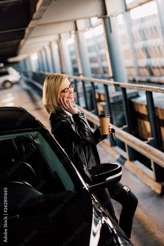 smiling blonde woman standing in parking with disposable cup while talking on smartphone near black automobile smiling blonde woman standing in parking with disposable cup while talking on smartphone near black automobile