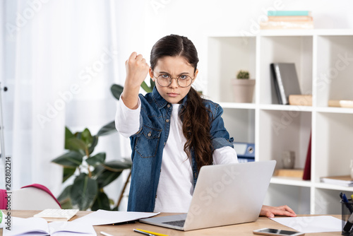 cute kid in glasses showing fist while standing near laptop in office