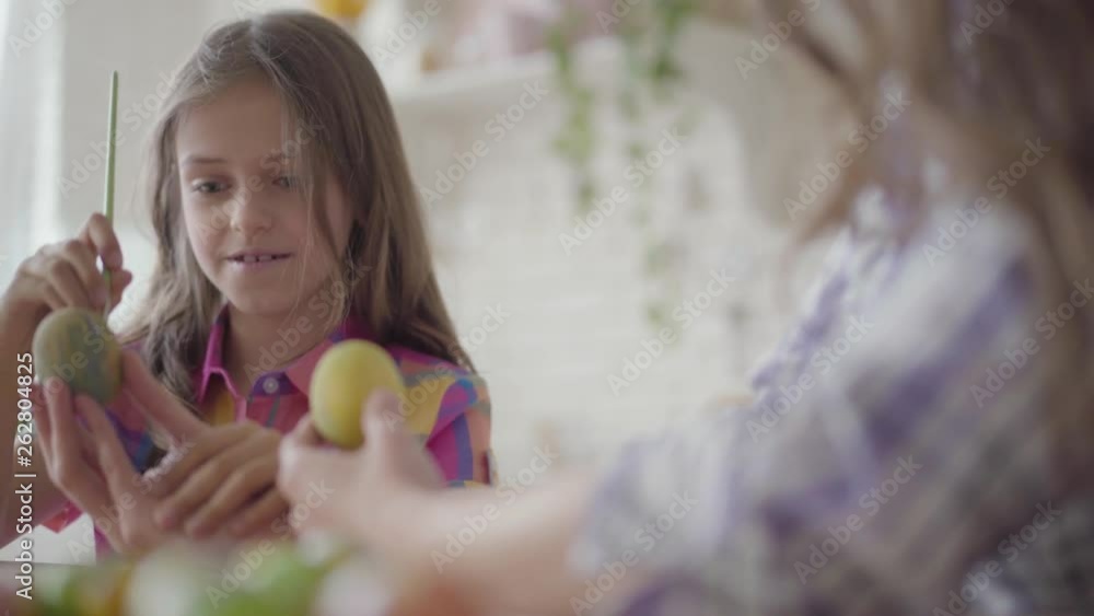 Female hand holding Easter egg while little smiling girl painting on it a heart with a small brush close up. Preparation for Easter holiday. Mother and daughter have fun together at the kitchen Female hand holding Easter egg while little smiling girl painting on it a heart with a small brush close up. Preparation for Easter holiday. Mother and daughter have fun together at the kitchen