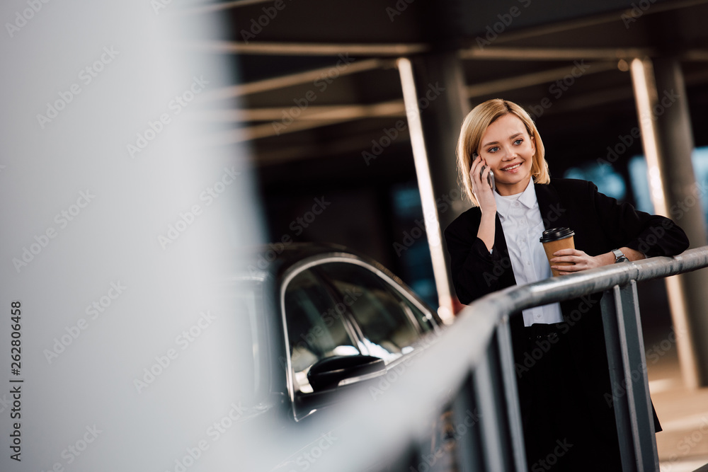 selective focus of cheerful blonde girl talking on smartphone and holding paper cup near black car selective focus of cheerful blonde girl talking on smartphone and holding paper cup near black car