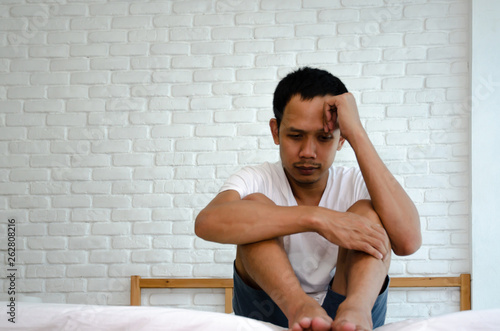 Men are sitting at the edge of the bed in the room and have stress.Young man wearing a white shirt with both hands on his head. Because of pain..