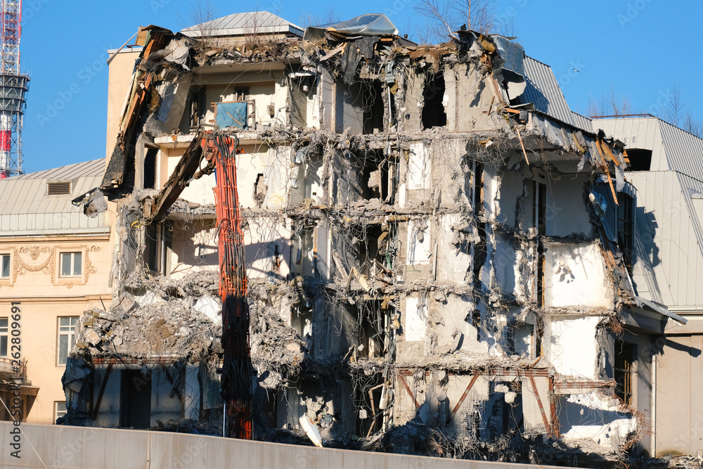 Building demolition with hydraulic excavator. Demolished destructed house ruins in european city on bright sunny day.