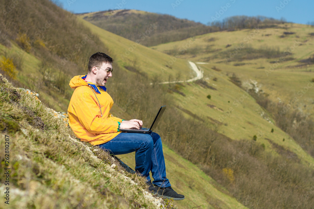 A tourist in a yellow jacket and glasses works on a laptop while on vacation in the mountains. In the background mountains. Freelance concept. Emotion of delight A tourist in a yellow jacket and glasses works on a laptop while on vacation in the mountains. In the background mountains. Freelance concept. Emotion of delight