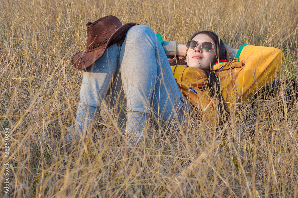 A young girl with long dark hair in a yellow jacket and glasses is lying in the yellow grass, leaning on a tourist backpack, her arms are thrown back over her head, and a leather cowboy hat on knee A young girl with long dark hair in a yellow jacket and glasses is lying in the yellow grass, leaning on a tourist backpack, her arms are thrown back over her head, and a leather cowboy hat on knee