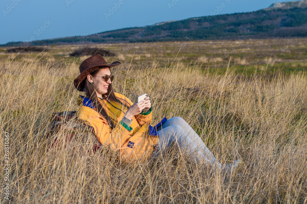 A young girl with long dark hair in a yellow jacket, a leather cowboy hat and glasses is sitting in the grass, leaning on a tourist backpack and making a photo on the phone. Background mountains, sky.
