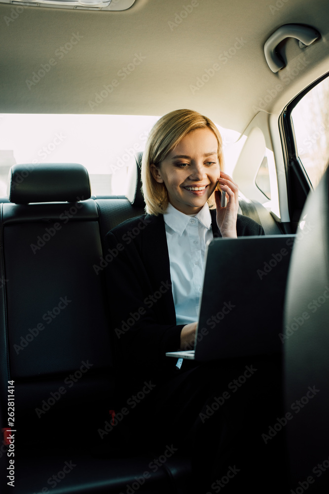 smiling blonde woman talking on smartphone and looking at laptop in car