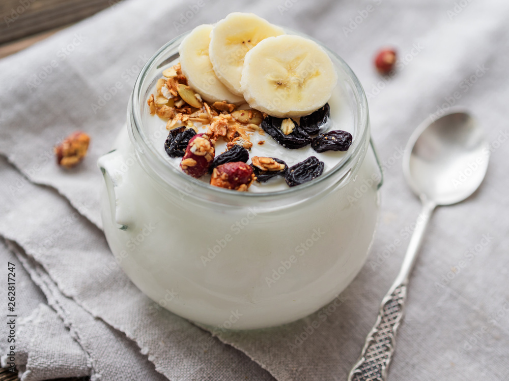 Natural homemade yogurt in a glass jar. Healthy food for breakfast with muesli. Jar with granola and banana slices on linen tablecloth on on wooden table. Natural homemade yogurt in a glass jar. Healthy food for breakfast with muesli. Jar with granola and banana slices on linen tablecloth on on wooden table.