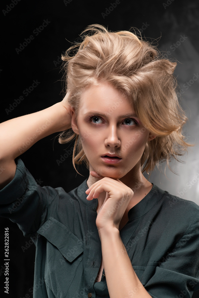 Beautiful young blonde girl with disheveled hairstyle and nude makeup, wearing a shirt and jeans emotionally posing on gray background. She is looking thoughtfully up and holding her head. Beautiful young blonde girl with disheveled hairstyle and nude makeup, wearing a shirt and jeans emotionally posing on gray background. She is looking thoughtfully up and holding her head.