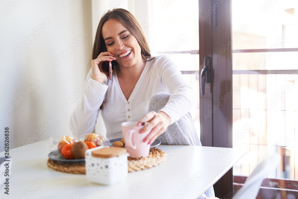 Attractive young happy woman using mobile phone and having breakfast at table near window at home Attractive young happy woman using mobile phone and having breakfast at table near window at home