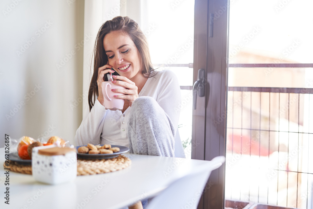 Attractive young happy woman using mobile phone and having breakfast at table near window at home Attractive young happy woman using mobile phone and having breakfast at table near window at home