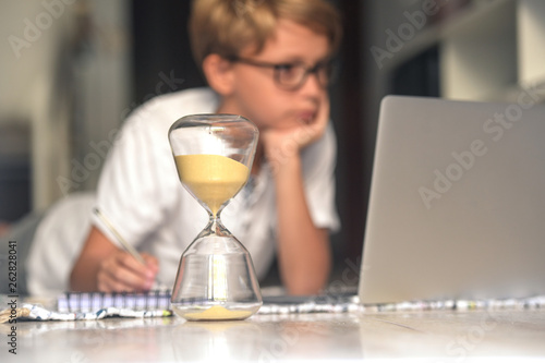 Handsome blond kid eyeglasses doing homework with computer keeping eye time available hourglass. healthy child searching information internet; Virtual classroom; education, home-schooling concept;