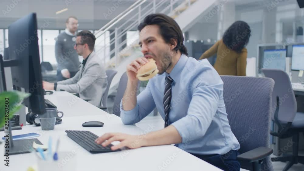 In the Bright Modern Office Young Businessman Eats Hamburger Sitting at His Desktop Computer, He Bites Angrily into the Bun and Continues to Work During His Lunch. In the Background Colleagues Working In the Bright Modern Office Young Businessman Eats Hamburger Sitting at His Desktop Computer, He Bites Angrily into the Bun and Continues to Work During His Lunch. In the Background Colleagues Working