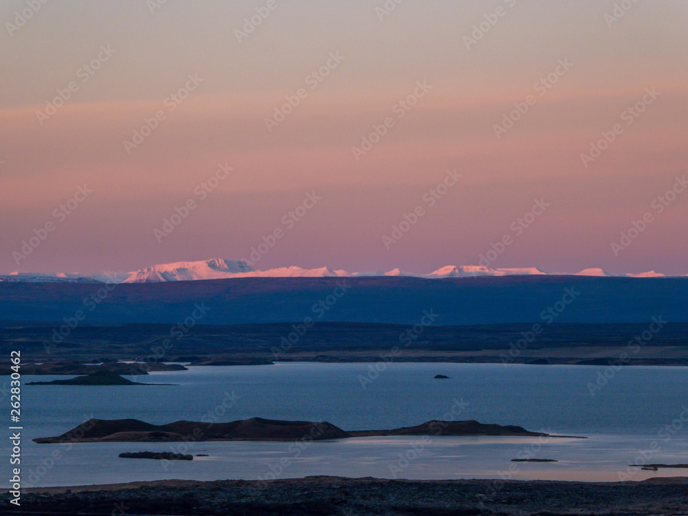 Sunrise over lava fields. Soft colors of the sky. High mountains in the back covered with snow. Lake in the middle, shines bright. Volcanic landscape, alienation and solitude. Harsh conditions.