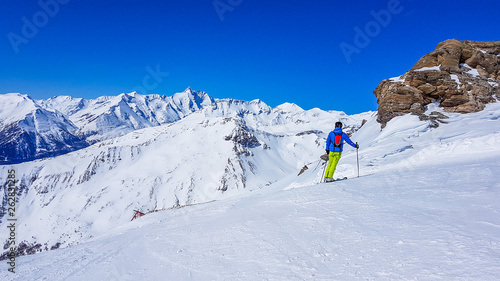 A skier standing on the side of the piste, enjoying the view. Perfectly groomed slopes. High mountains surrounding the man wearing yellow trousers and blue jacket. Man wears helm for the protection.