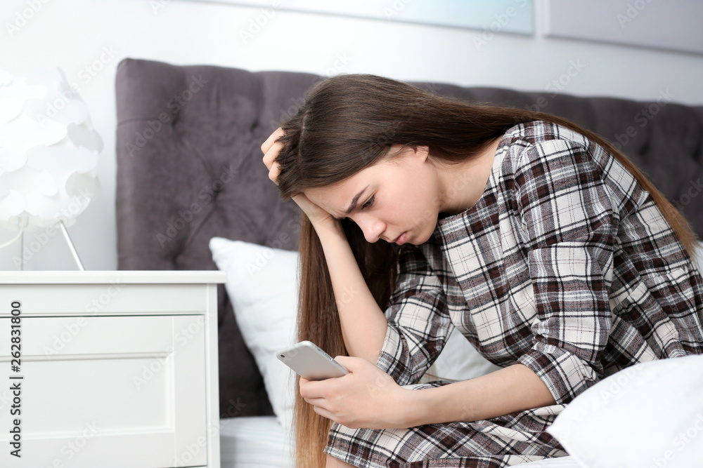 Upset teenage girl with smartphone sitting on bed. Space for text Upset teenage girl with smartphone sitting on bed. Space for text