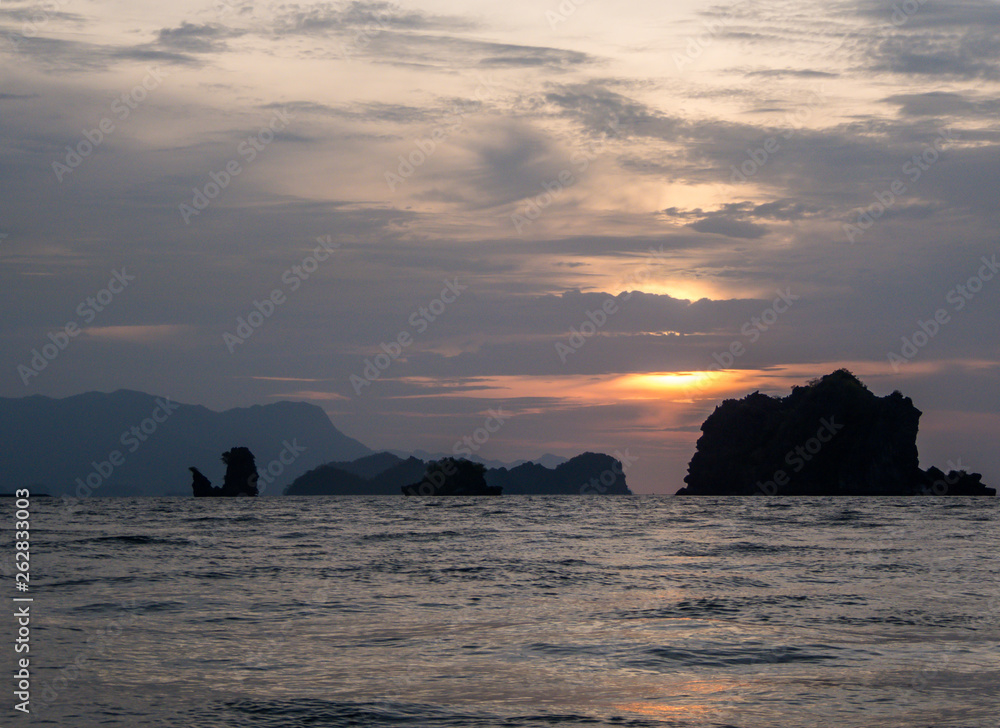 Sunset over the sea. Sun goes down behind the clouds. Sunbeams reflecting in the calm surface of the sea. A small rocky formation in the middle of the sea. In the back taller mountains are emerging.