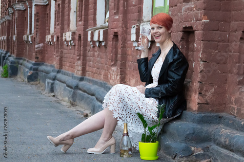 Young red-haired woman in a white dress sitting on the street and drinking white wine