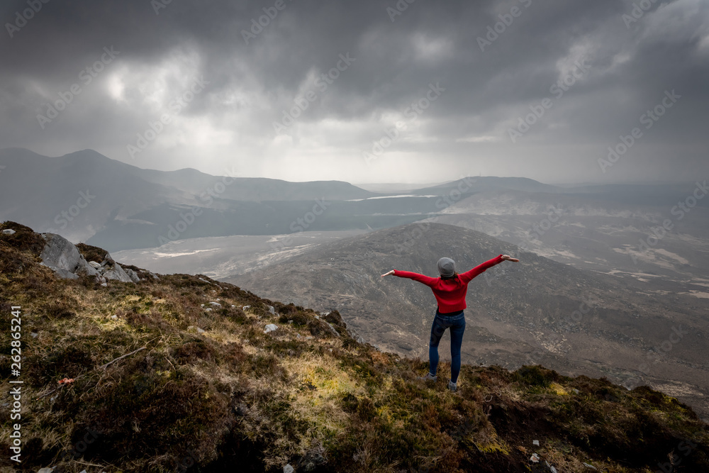 une femme écarte les bras en haut d'une montagne devant un paysage grandioses  sous un ciel menaçant