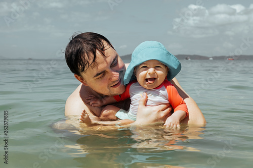 Child shows tongue, she is enjoy. Happy laughing infant baby with dad having fun in the sea. Father teaches daughter to swim and water. photo