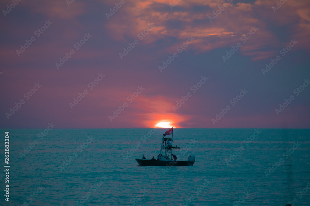 A boat on the teal green ocean water in front of the setting sun amid the blue and purple clouds of evening, as seen from a beach on the Gulf of Mexico near Englewood, Florida, USA, in early spring