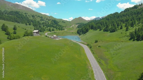 AERIAL: Tourists on motorbikes ride along the scenic road towards the gorgeous emerald lake. Flying above an asphalt road running through the picturesque green valley and towards the rocky mountains.